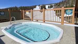 Een hottub met een houten terras en leuning, omringd door een besneeuwd landschap en bomen.