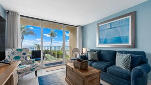 A living room with a blue sofa, a wooden coffee table, and a large window with a view of palm trees and the ocean.