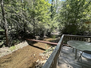 A wooden deck with a table and chairs, overlooking a forested area with a stream.