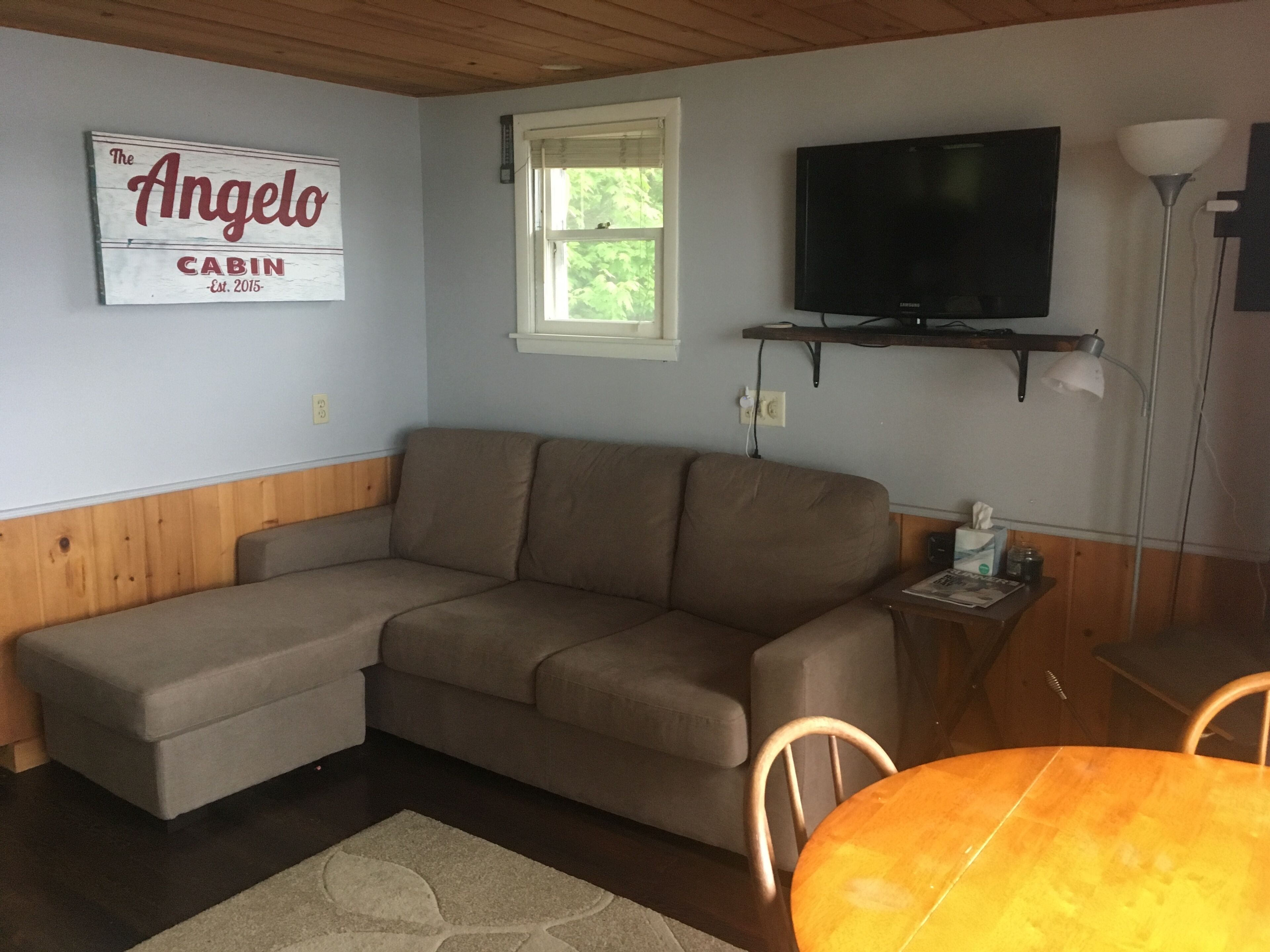 A living room with a sectional sofa, a flat-screen TV mounted on the wall, a small table with a lamp, and a sign that reads "The Angelo Cabin."