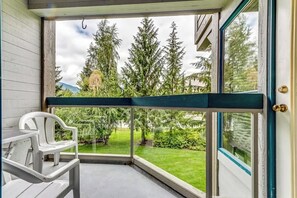 Balcón en esquina con vistas a un césped verde y árboles, una silla de plástico blanca y una mesa.