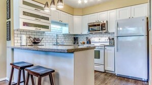 A kitchen with white cabinets, a granite countertop, a stainless steel refrigerator, and a built-in oven. There are two bar stools and pendant lights above the counter. - Greystone Lodge 210 Ski-in/Out by Acer Vacations (Whistler)