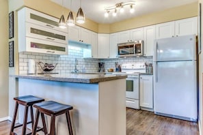 A kitchen with white cabinets, a granite countertop, a stainless steel refrigerator, and a built-in oven. There are two bar stools and pendant lights above the counter. - Greystone Lodge 210 Ski-in/Out by Acer Vacations (Whistler)