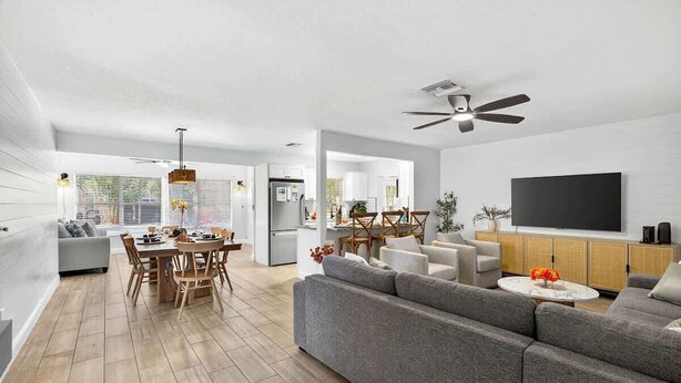 A modern living room with a grey sofa, dining table, and chairs, a TV on a wooden cabinet, and a ceiling fan. - Lantana Cabana 3BR North Clearwater Beach Home (Cleawater)