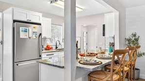 A modern kitchen with white cabinetry, a stainless steel refrigerator, and a dining area with wooden chairs and a marble countertop. - Lantana Cabana 3BR North Clearwater Beach Home (Cleawater)