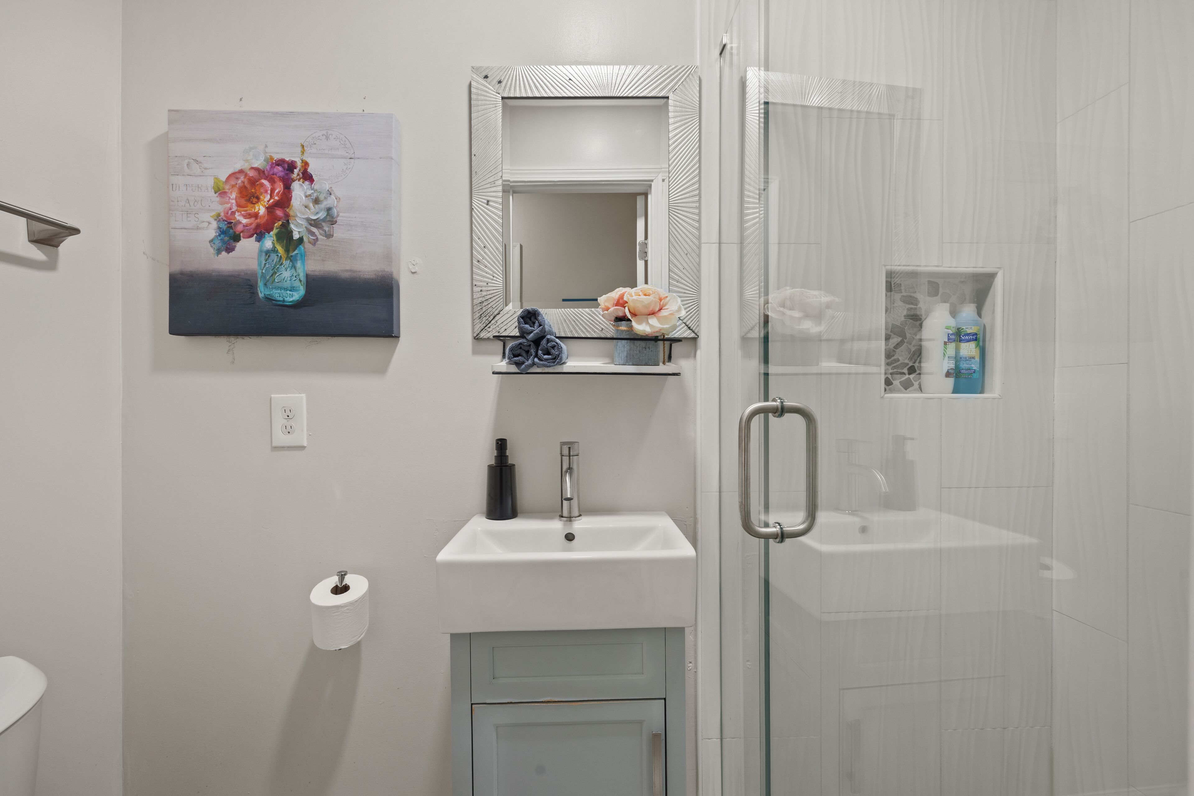 Une salle de bain avec un lavabo blanc, un miroir, une douche en verre et un tableau de fleurs accroché au mur.