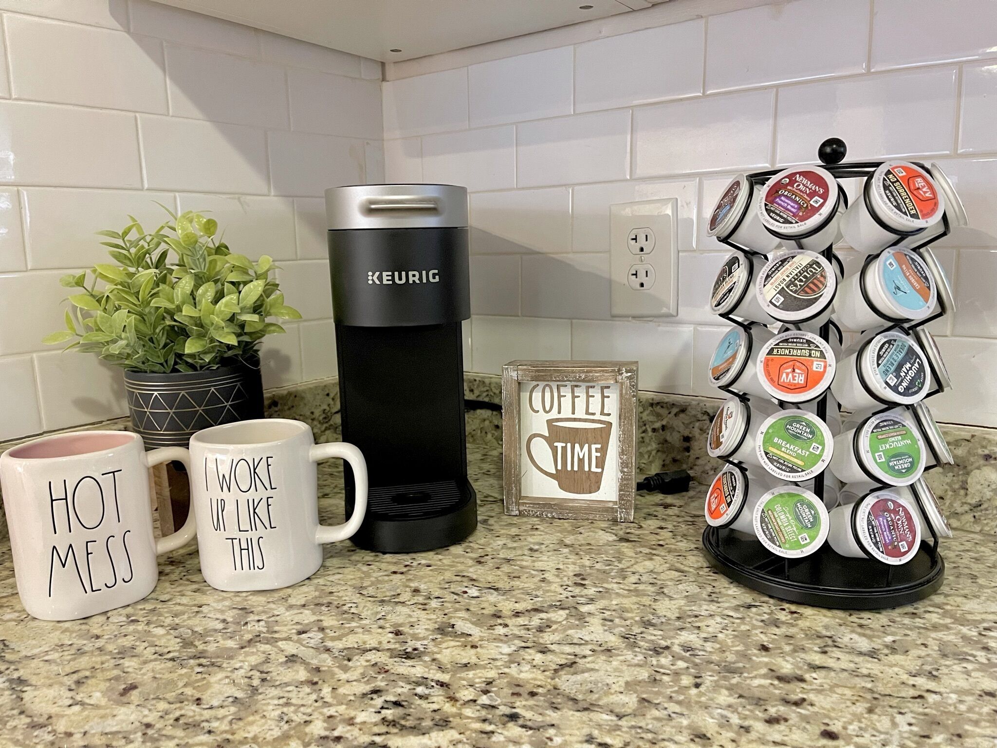 Una encimera de cocina con una cafetera Keurig, dos tazas con texto, un portalápices para cápsulas de café y un pequeño cartel que dice 'Hora de café'.