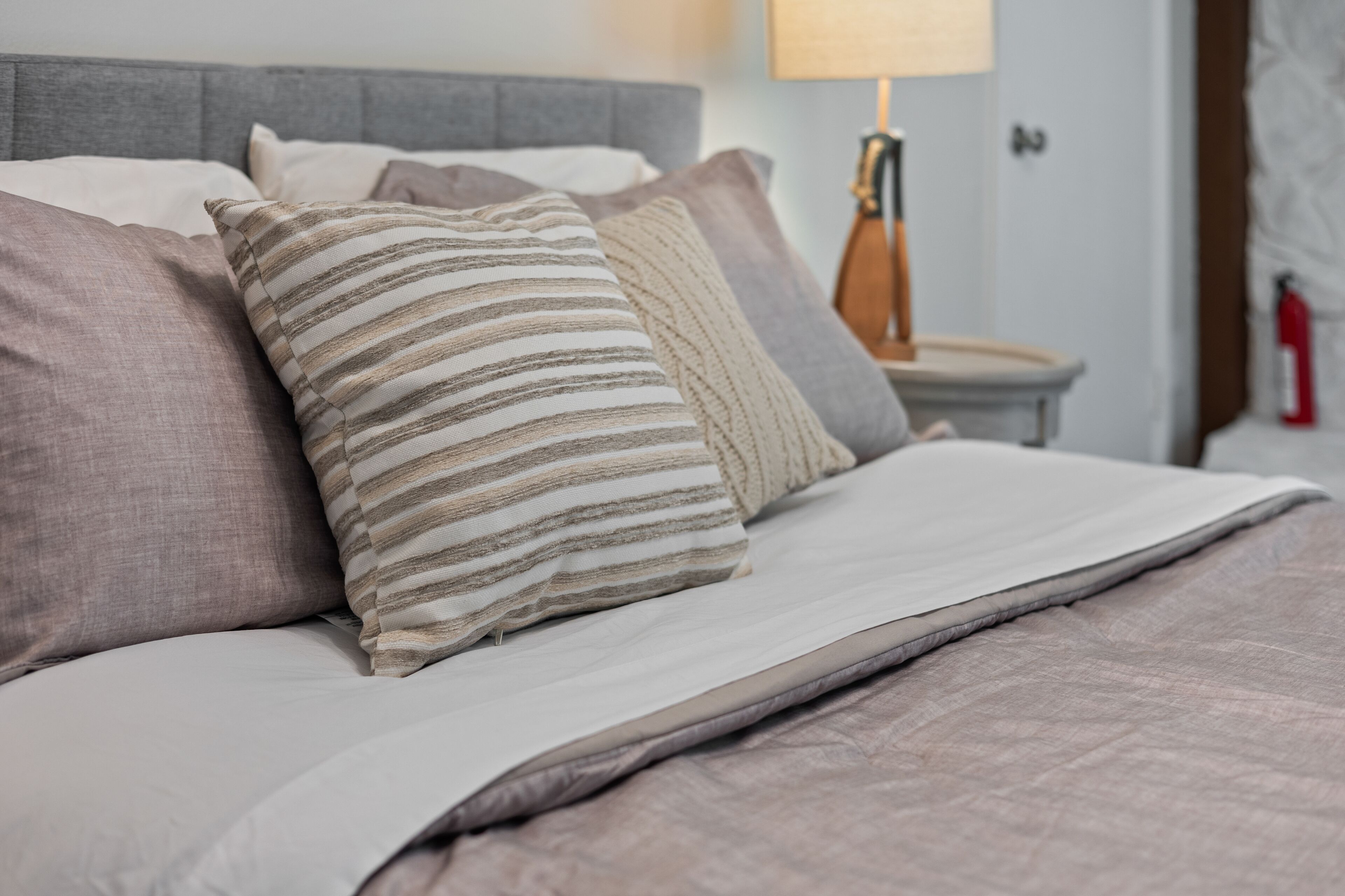 A neatly made bed with striped and solid pillows, a grey headboard, and a bedside table with a lamp.