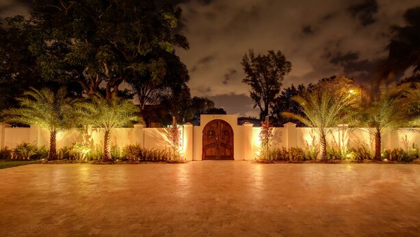 Une vue nocturne de l’entrée d’un bâtiment, avec une grande porte en bois, entourée de palmiers et éclairée par une lumière chaleureuse.