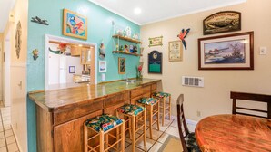 A kitchen with a wooden bar and stools, a dining table, and various wall decorations including a framed picture of a ship.