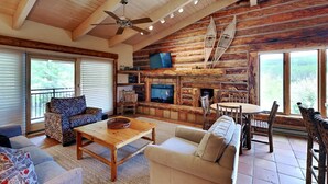 A cozy cabin interior with a fireplace, wooden walls, a ceiling fan, and a large window. - 🌲Timberline A3E Deluxe 2-Bedroom + Loft ~ 2/bath ~Hot Tub~Pool~WiFi~Ski-in/out~Local Shuttle🌲 (Snowmass Village)
