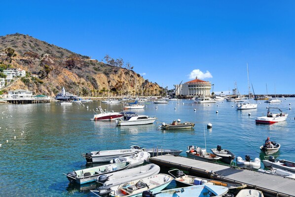 Boats docked in a serene marina, surrounded by a hill with lush vegetation and a unique building in the distance.