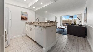 A modern kitchen with white cabinetry and a granite countertop, leading to a dining area with a dark sofa and a view of the outdoors.