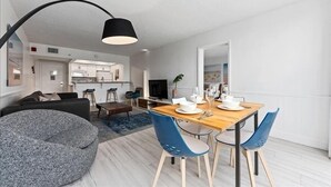 A modern living room with a dining area, a kitchen in the background, and a large window.