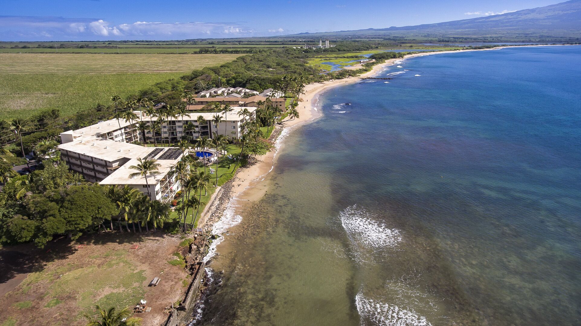 An aerial view of a coastal hotel with a beach, clear waters, and a mountainous backdrop.