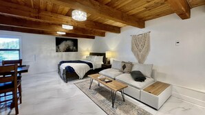 A modern bedroom with a wooden ceiling, a bed, a sofa, a coffee table, and a dining area with chairs. - Waterfront | Hot Tub | Firepit | King Bed - 6H (Ucluelet)