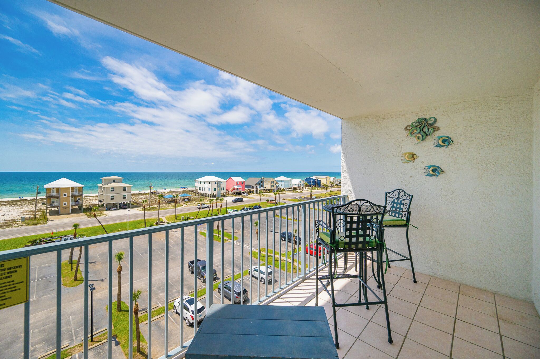 A balcony with a view of the beach, a parking lot, and a building with a red roof.