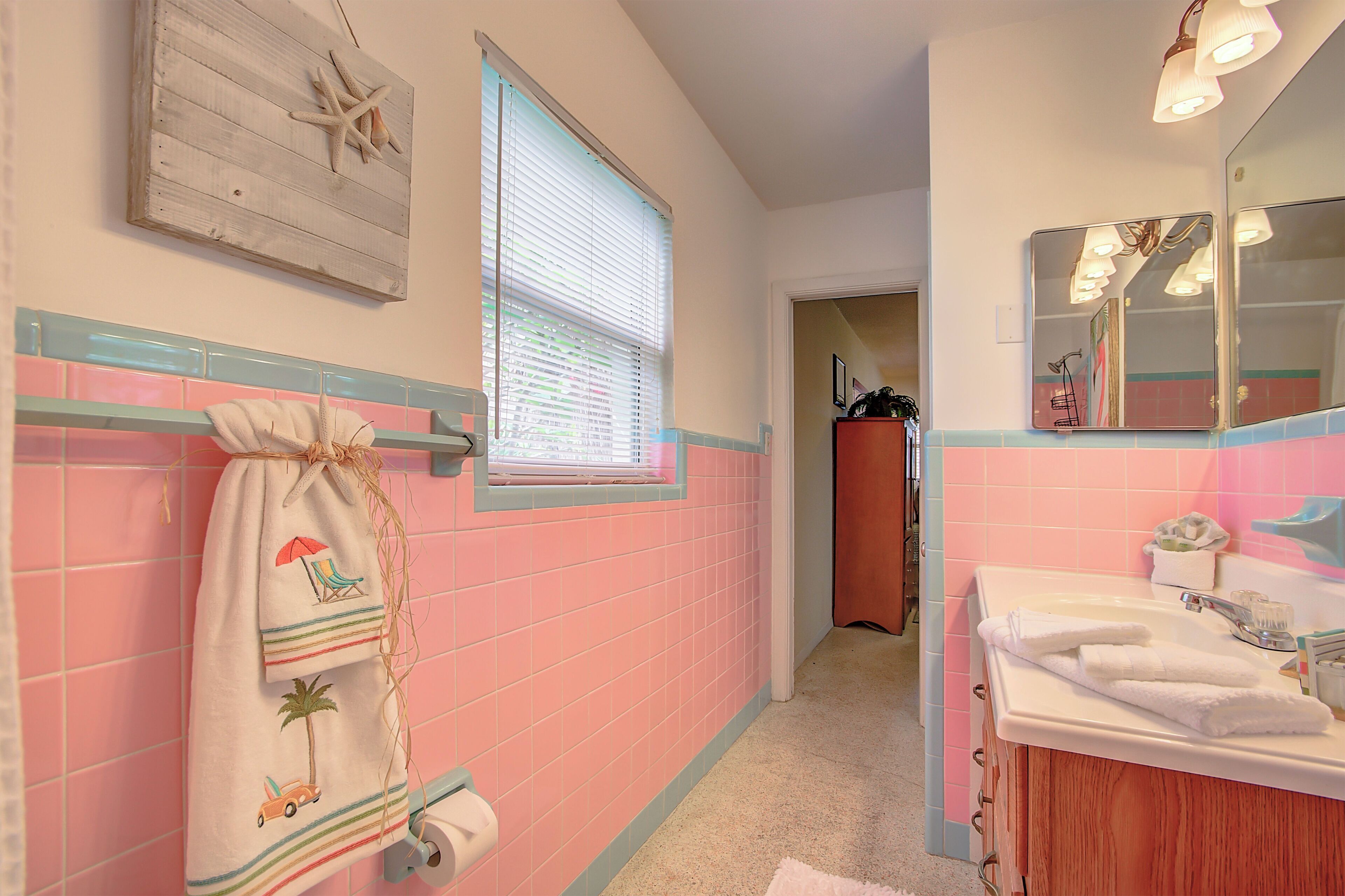 Un baño con azulejos rosas, una ventana con persianas, un perchero con una toalla de temática playera y un lavabo con espejo.
