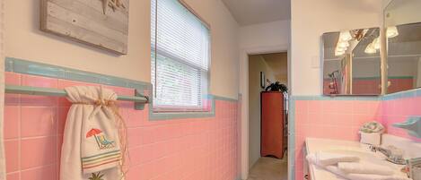 Un baño con azulejos rosas, una ventana con persianas, un perchero con una toalla de temática playera y un lavabo con espejo.