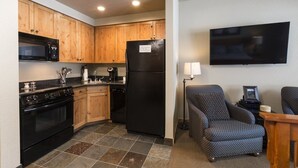 A compact kitchen with wooden cabinets, a black refrigerator, a microwave, and a stove. There is a flat-screen TV mounted on the wall and a chair in the living area. - Studio Style Lodging Ski-In/Ski-Out (Whitefish)