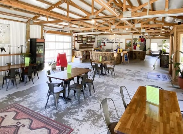 Un vaste espace intérieur doté de hauts plafonds et de poutres apparentes. On y trouve plusieurs tables et chaises disposées pour les repas ou les réunions. Un comptoir où sont présentés divers articles, ainsi qu'un présentoir de vêtements, sont visibles au fond.