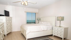 Ein Schlafzimmer mit Bett, Schminktisch, Nachttisch, Deckenventilator und Meerblick durch das Fenster.