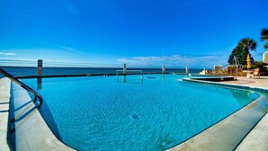 A clear blue pool with a view of the ocean, surrounded by a deck with lounge chairs and a palm tree.