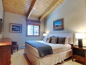 A bedroom with a large bed, two bedside lamps, a wooden desk, a chair, and two framed pictures on the walls.