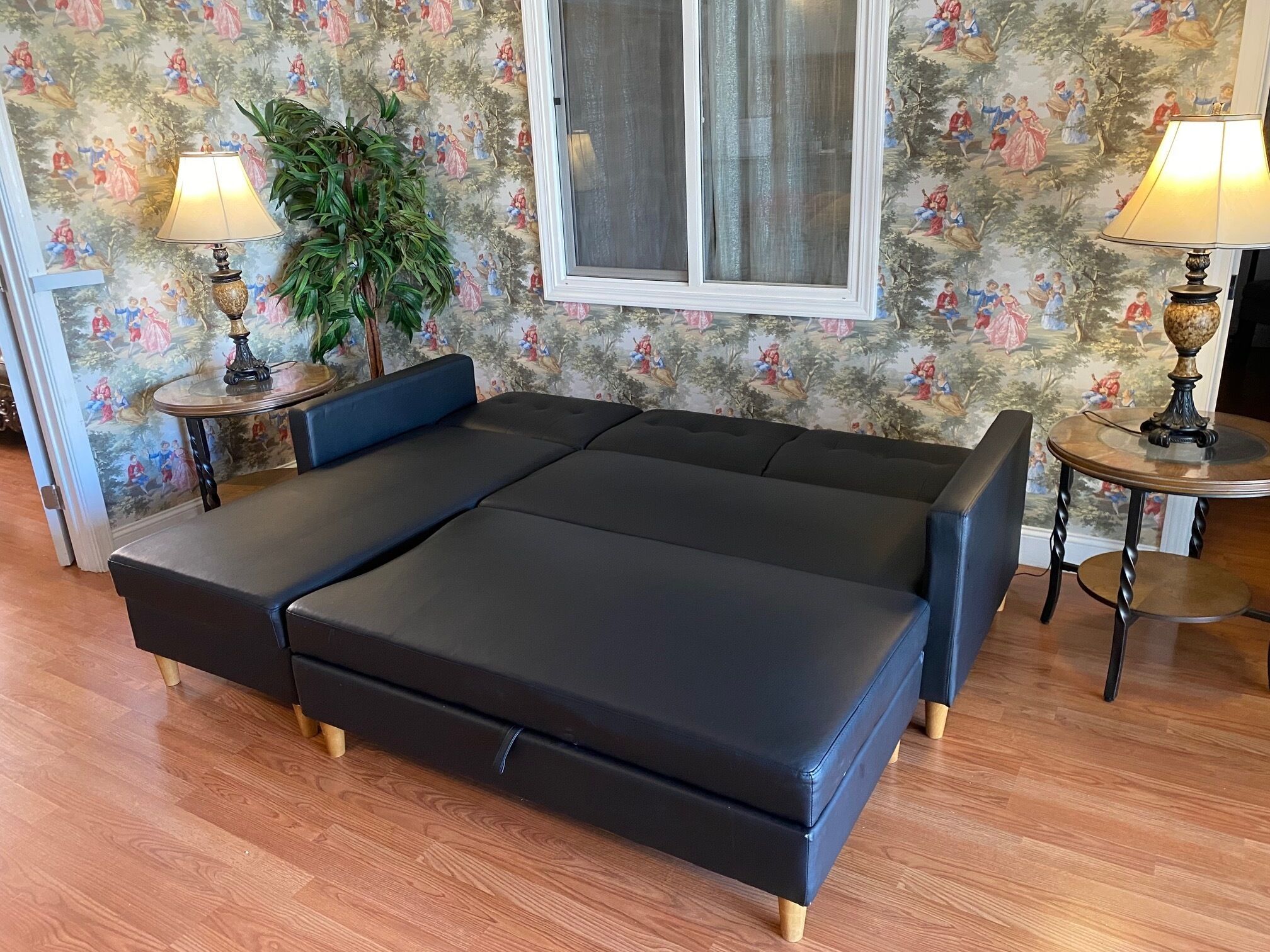Een kamer met een zwart slaapbank, twee kleine ronde tafels met lampen, een potplant en een raam met gordijnen.