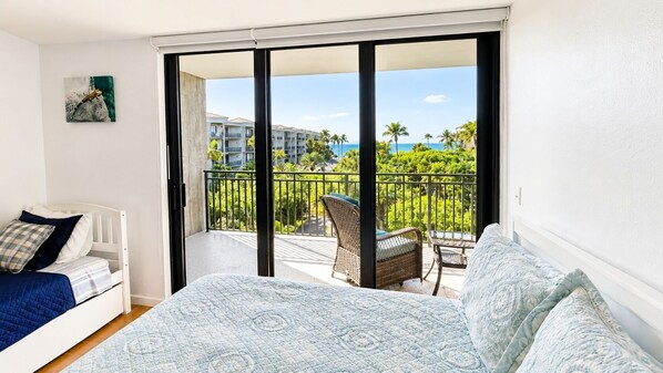 A bedroom with a bed, a chair, and a balcony with a view of buildings and palm trees.