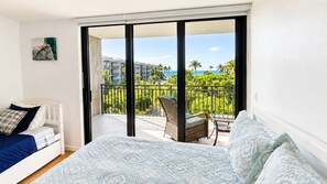 A bedroom with a bed, a chair, and a balcony with a view of buildings and palm trees.