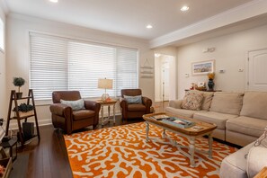 Uma sala de estar com um sofá de couro marrom, um sofá bege e um tapete estampado. Há duas poltronas, uma mesa de centro e uma mesa lateral pequena com uma luminária.