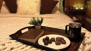A cozy bedroom with a bed, pillows, a bedside table with a lamp, a tray with chocolates and a mug, and a book.