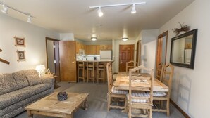Ein gemütliches Wohnzimmer mit einer Couch, einem Holztisch und einem Esstisch mit Stühlen. Im Hintergrund ist eine Küche mit Holzschränken und einem Kühlschrank zu sehen.