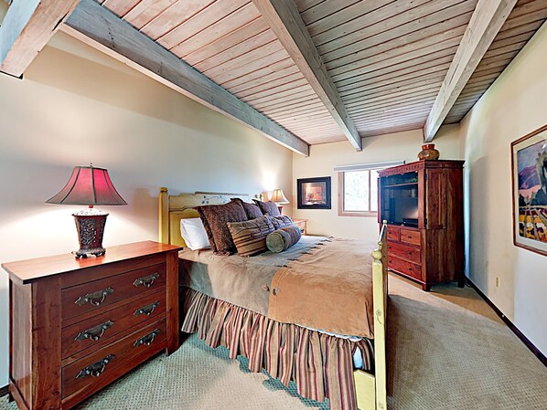 A bedroom with a wooden ceiling, a bed with patterned bedding, a nightstand with a lamp, a dresser, and a cabinet.