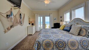 A bedroom with a bed, a television, a ceiling fan, a dresser, a chair, and a view of the ocean through a door. - Five Bedroom Oceanfront Home - Just Chilln (North Topsail Beach)