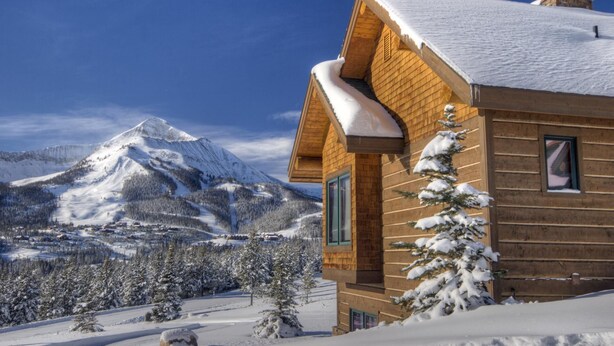 A wooden cabin with a snow-covered roof, surrounded by a snowy landscape and a mountain in the background.