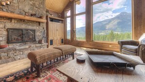 A cozy room with a stone fireplace, wooden beams, and large windows offering a mountain view.