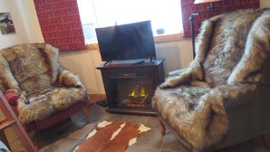 Una habitación con dos sillones cubiertos de piel, un televisor sobre un mueble bajo, una chimenea con leña y una cortina roja estampada.