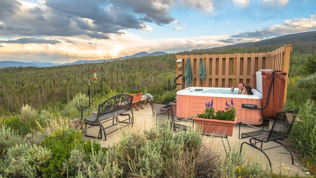 Un jacuzzi rodeado de asientos al aire libre y una terraza de madera, con un bosque y montañas como fondo.