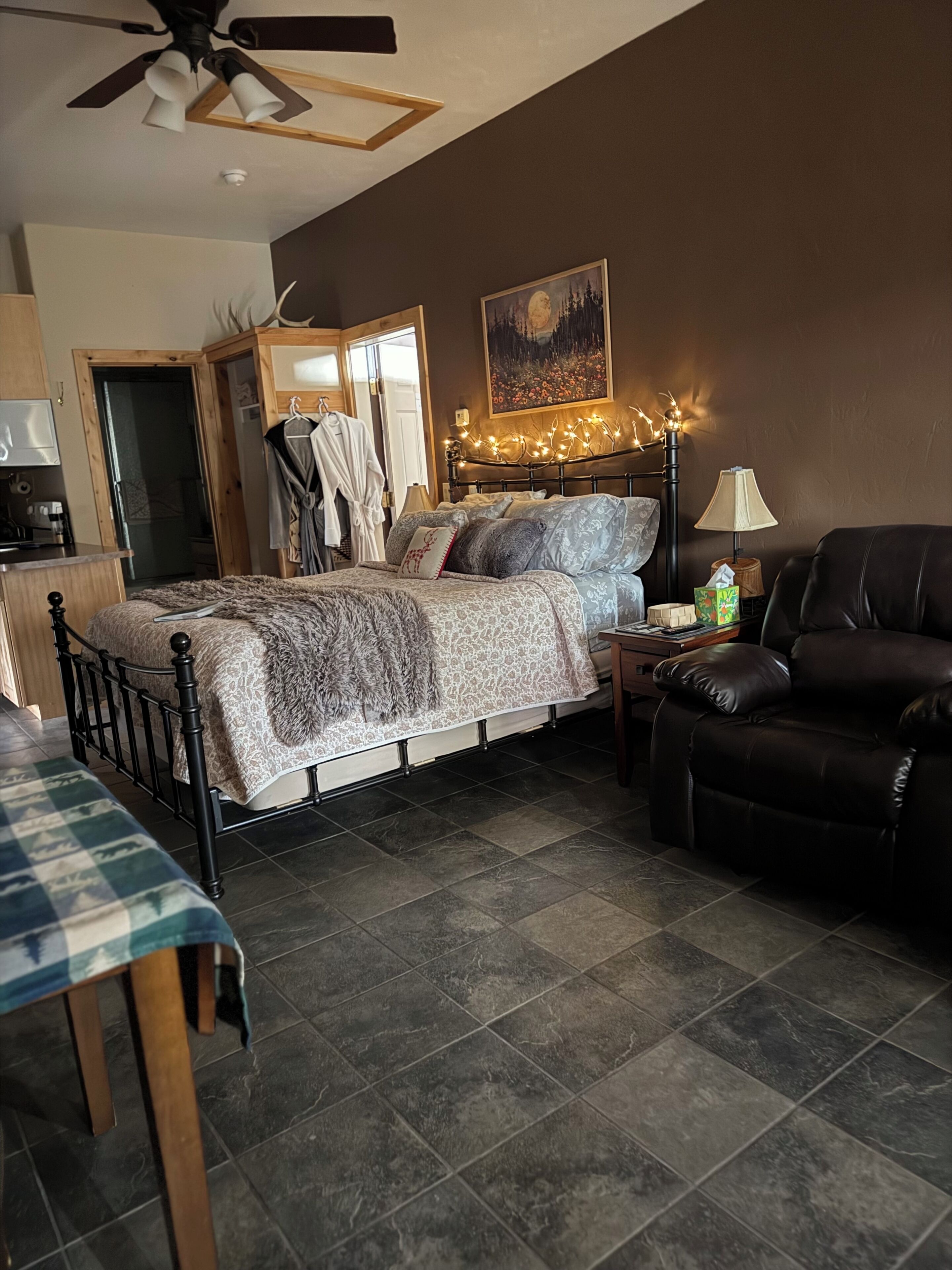 Une chambre à coucher comprenant un lit, une chaise, un bureau, un ventilateur de plafond, un tableau et une table de chevet avec une lampe.
