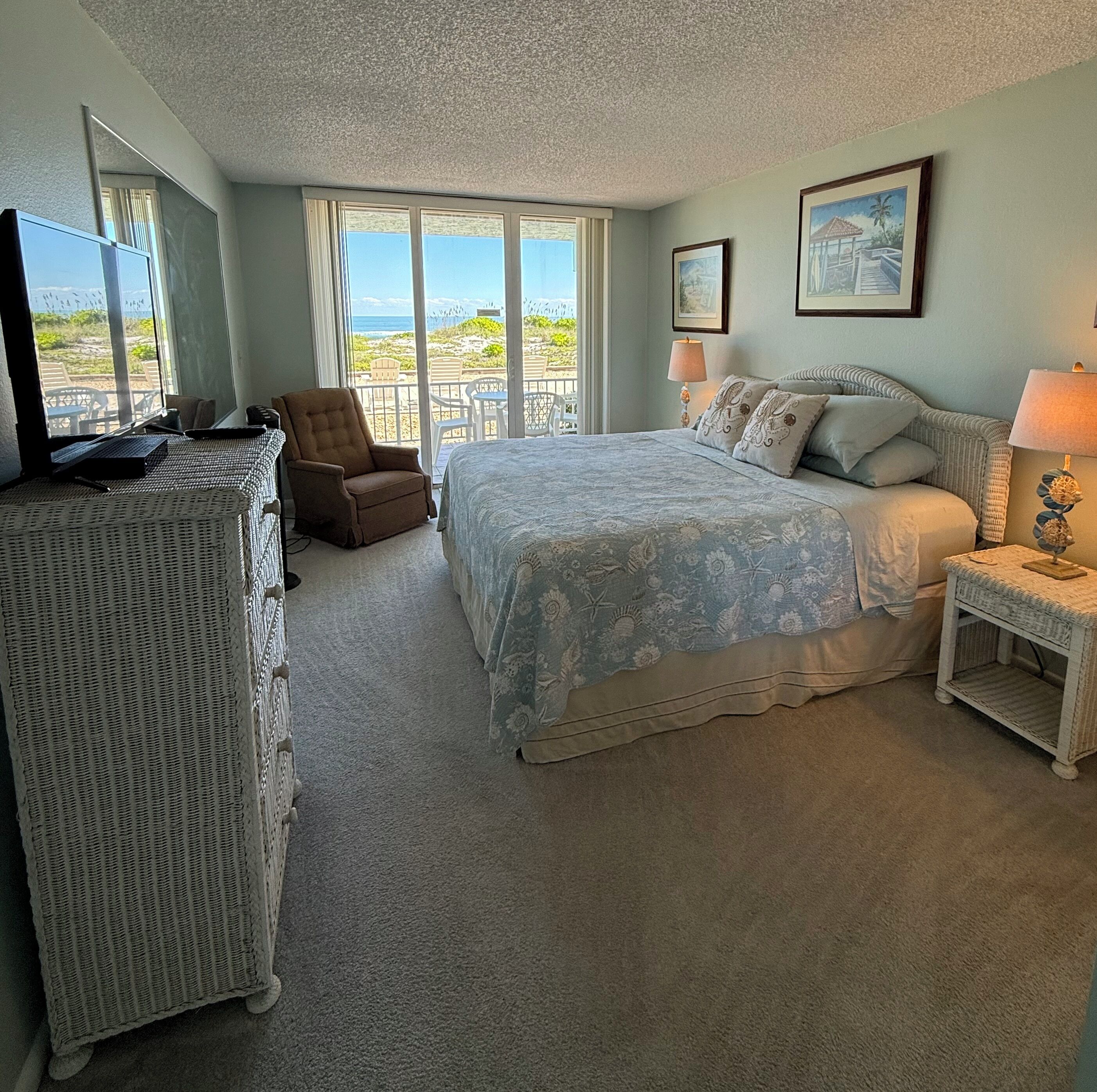 A bedroom with a large bed, a chair, a desk, and a balcony with a view of the beach.