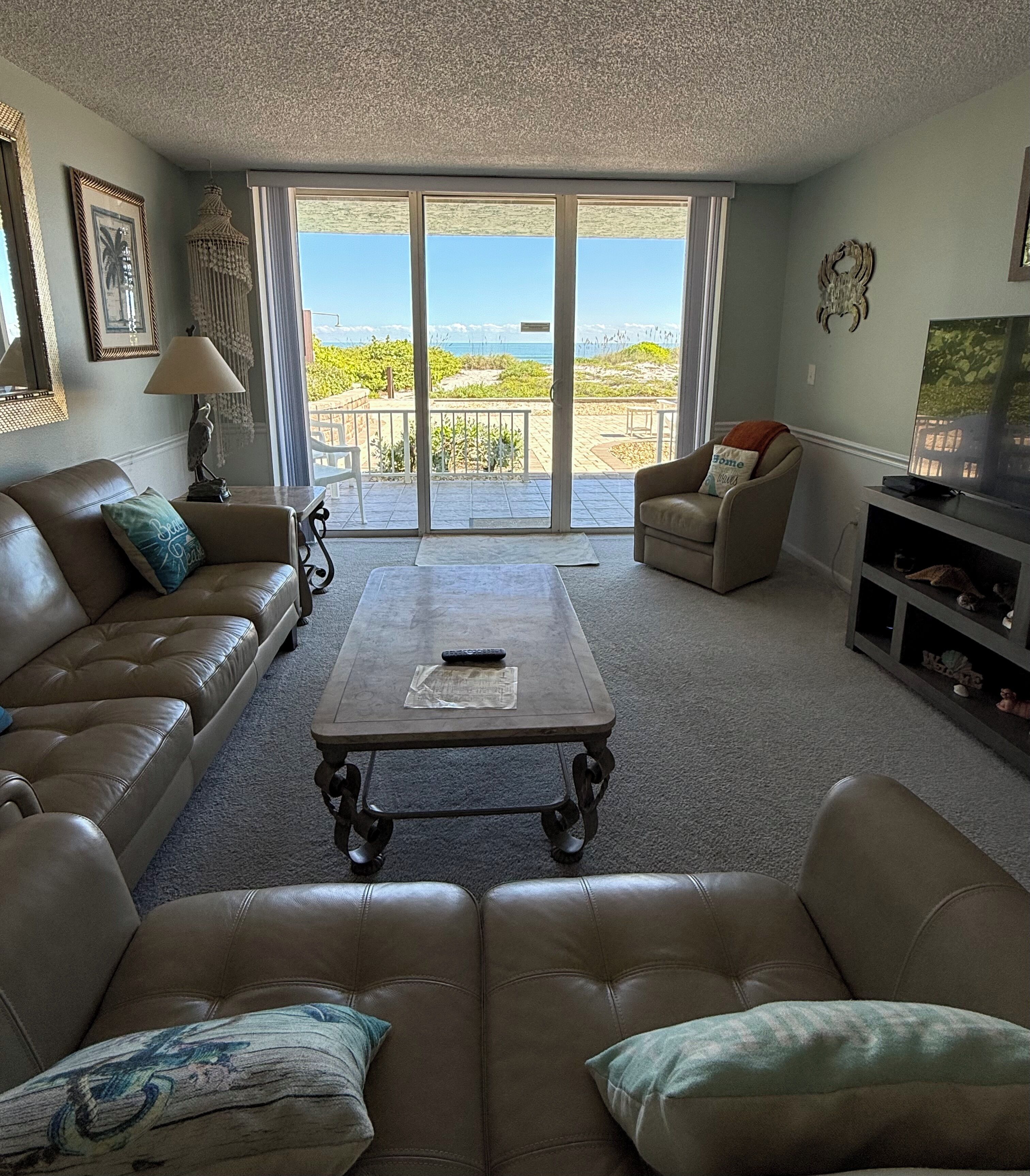 Un salon avec un canapé, une table basse et un téléviseur. Il y a un balcon avec vue sur la plage.