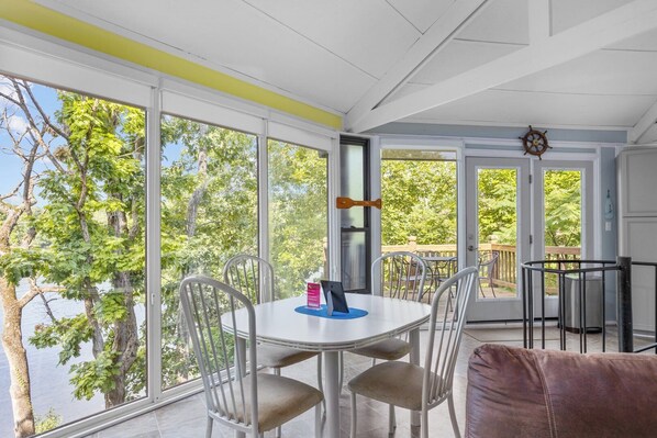 A sunlit room with large windows, a round table, and chairs, overlooking a deck and trees.