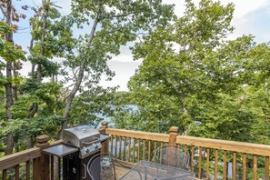 A deck with a grill, chairs, and a view of a lake surrounded by trees.
