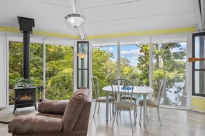 A room with a fireplace, a sofa, a dining table with chairs, and large windows offering a view of trees and a body of water.