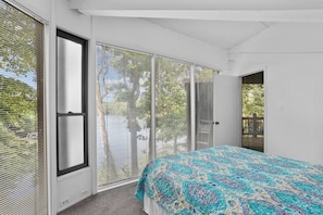 Un dormitorio con cama, ventanas amplias con persianas y vista a árboles.