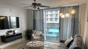 A modern living room with a flat-screen TV, a ceiling fan, a sofa, a patterned ottoman, and a sliding glass door leading to a balcony. - Breathtaking 2/2 Ocean View Fully Remodeled Condo! (Hollywood)