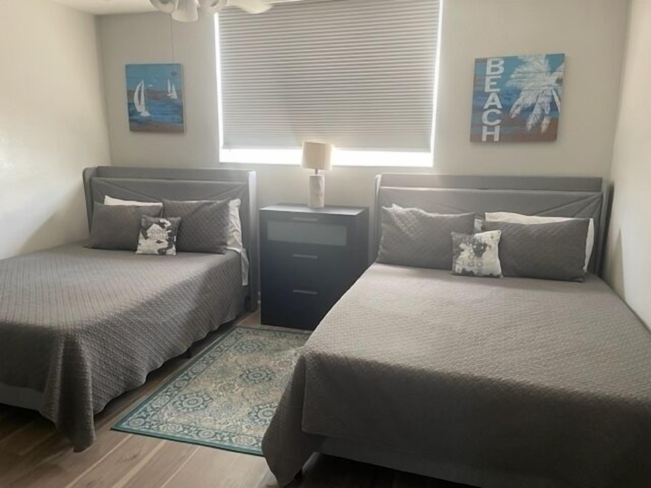 A bedroom with two single beds, a nightstand, a window with blinds, and wall art.