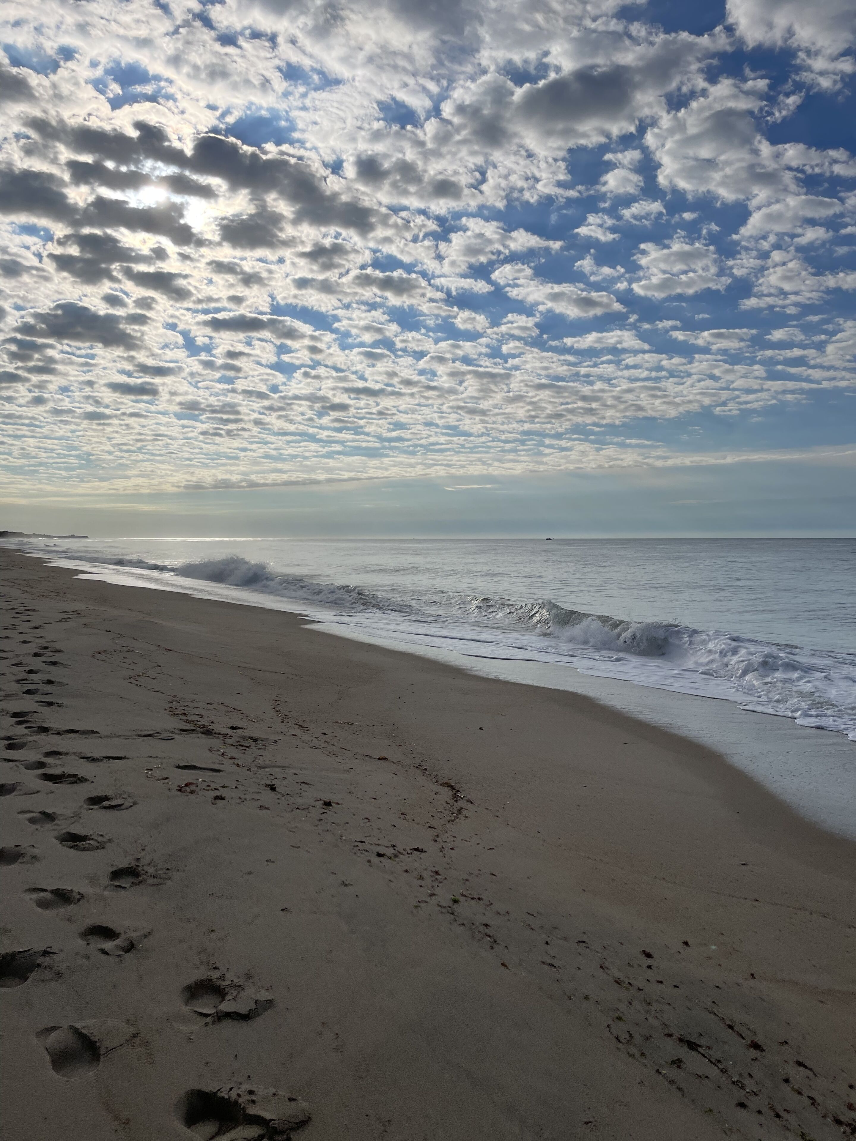 En strand med fodspor, der fører ned mod vandet, bølger der forsigtigt slår ind mod kysten, og en delvist overskyet himmel.